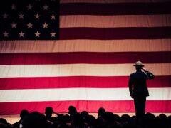 man standing on stage facing an american flag